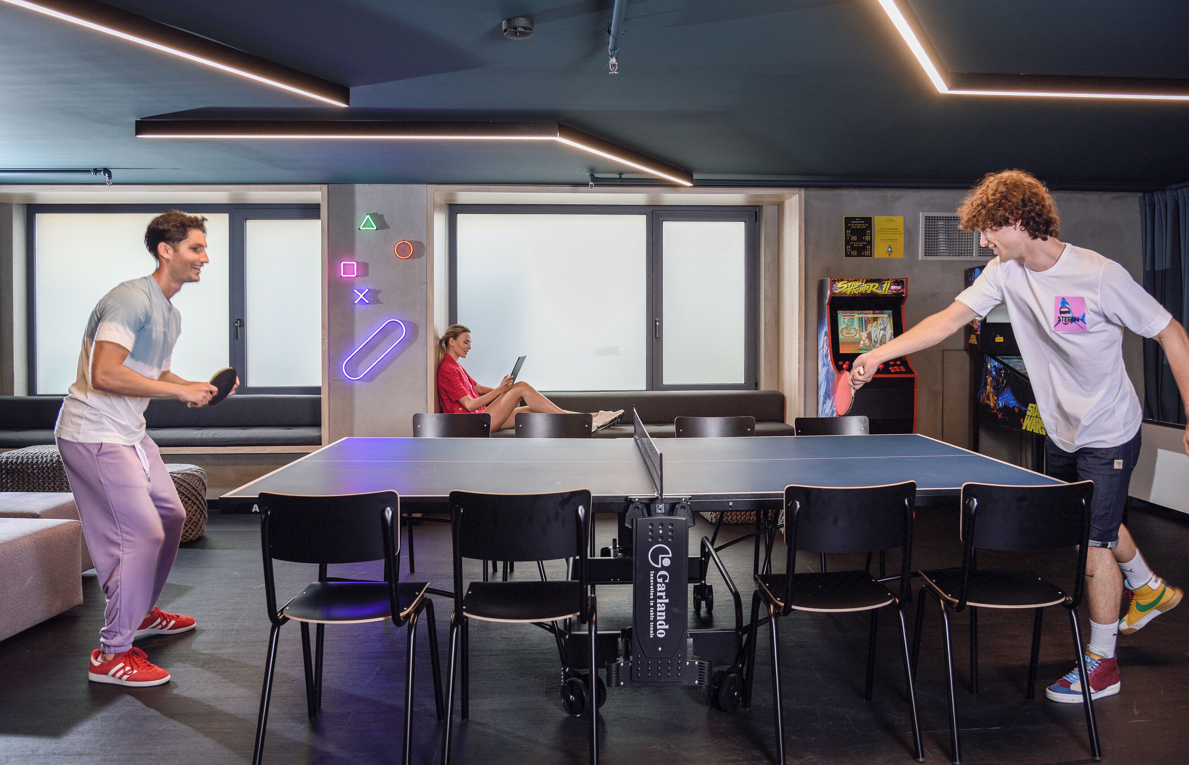 A ping pong table with chairs, a sofa, and a neon sign.