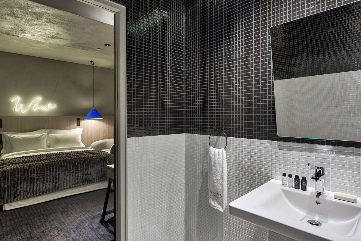 A modern hotel room with a bed, a neon 'Wow' sign, a blue pendant light, a white sink with toiletries, and a large mirror.