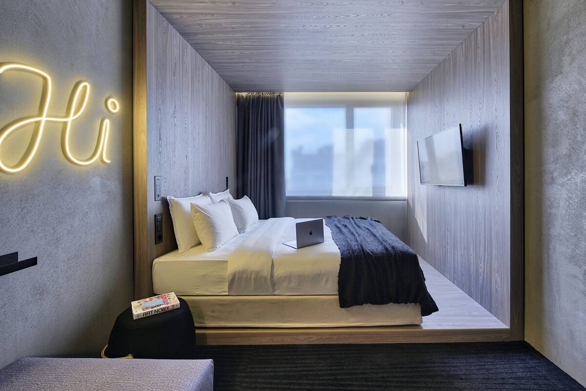 A modern hotel room with a bed, a desk, a laptop, a TV, and a neon sign.