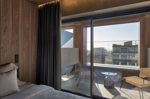 A modern hotel room with a large sliding glass door leading to a balcony, a bed with white linens, and a wicker chair.