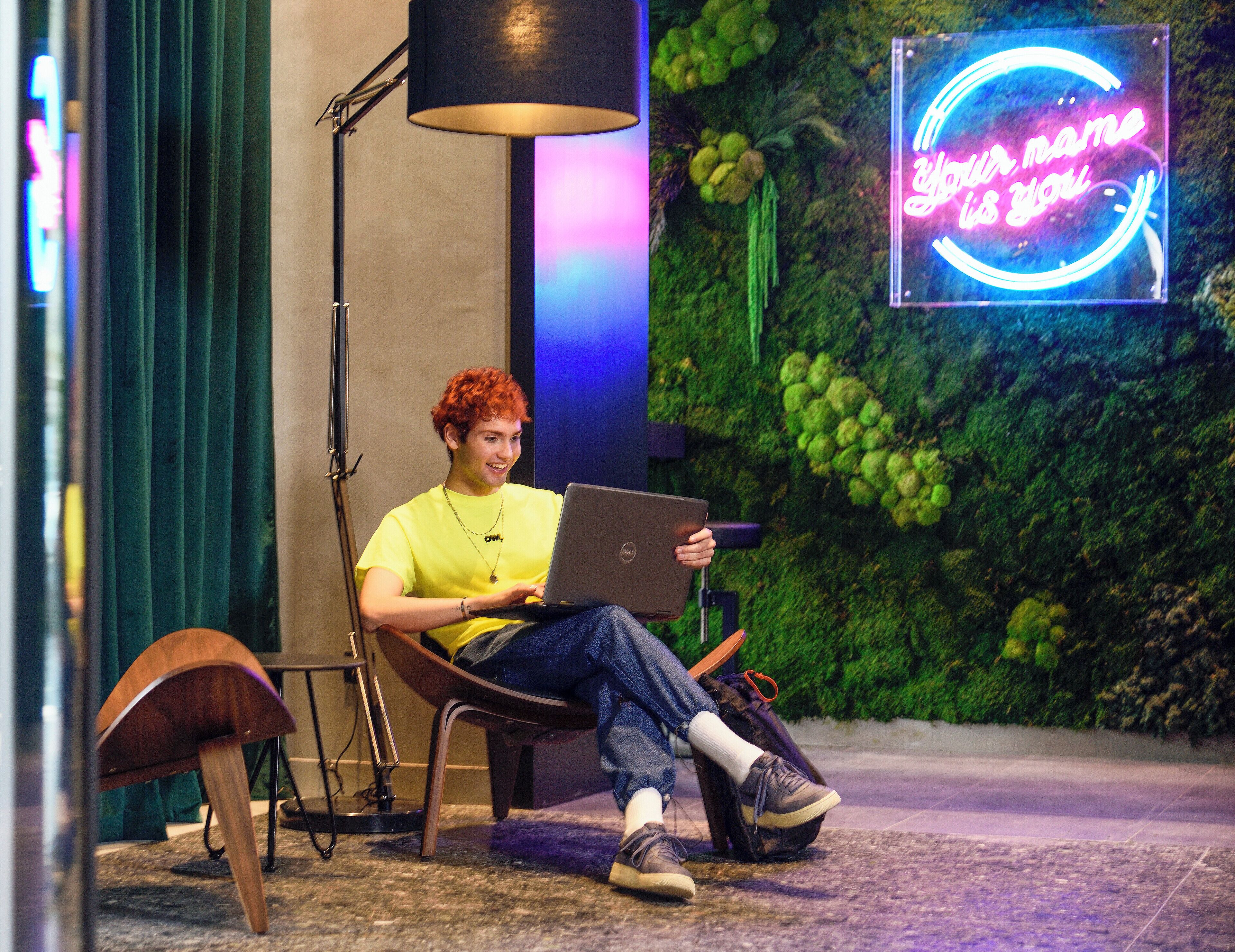 A person sitting on a chair with a laptop, a guitar, and a backpack on the floor.