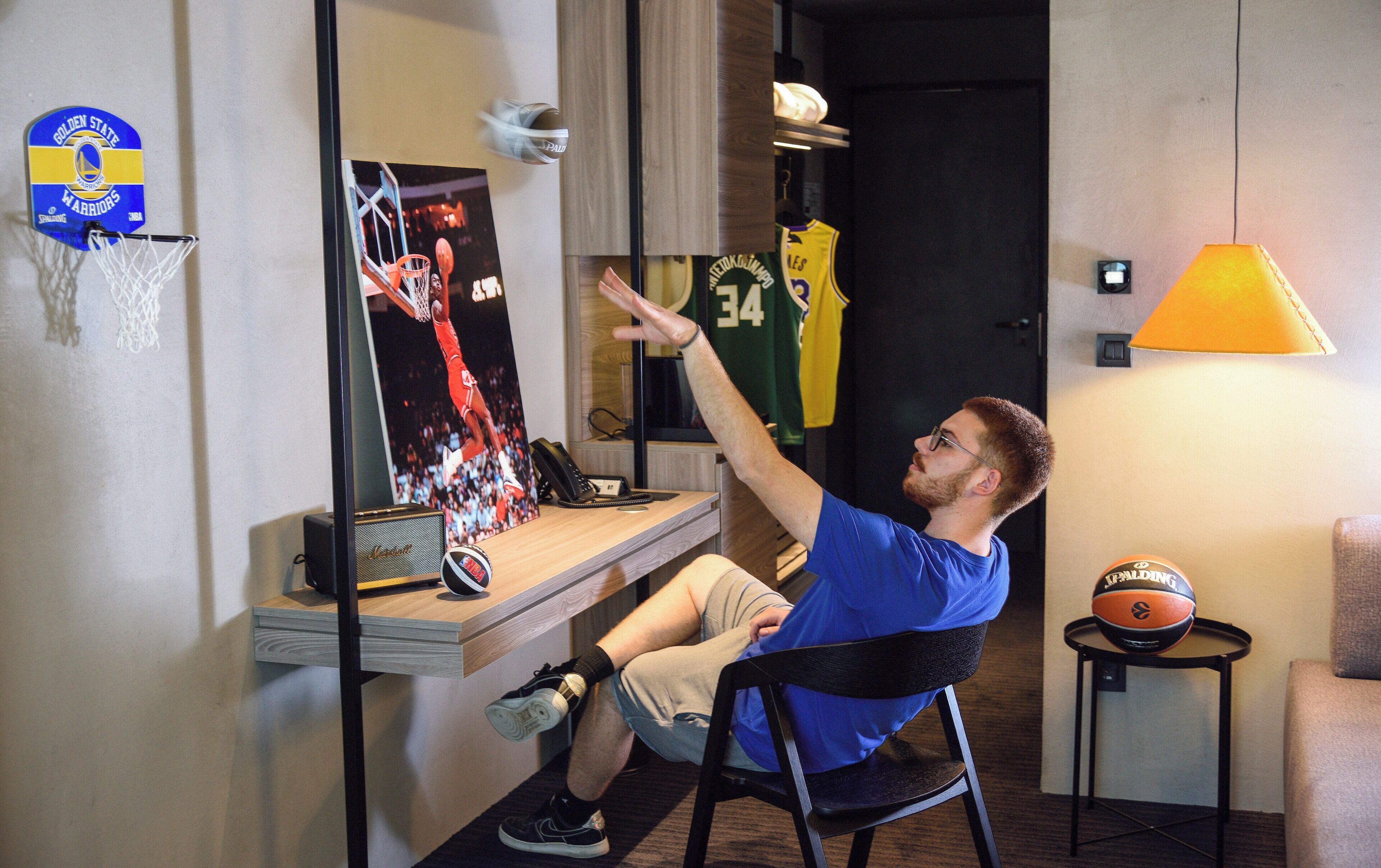 A room with a basketball hoop, a poster, a desk with a computer, a chair, a lamp, and a basketball on a side table.