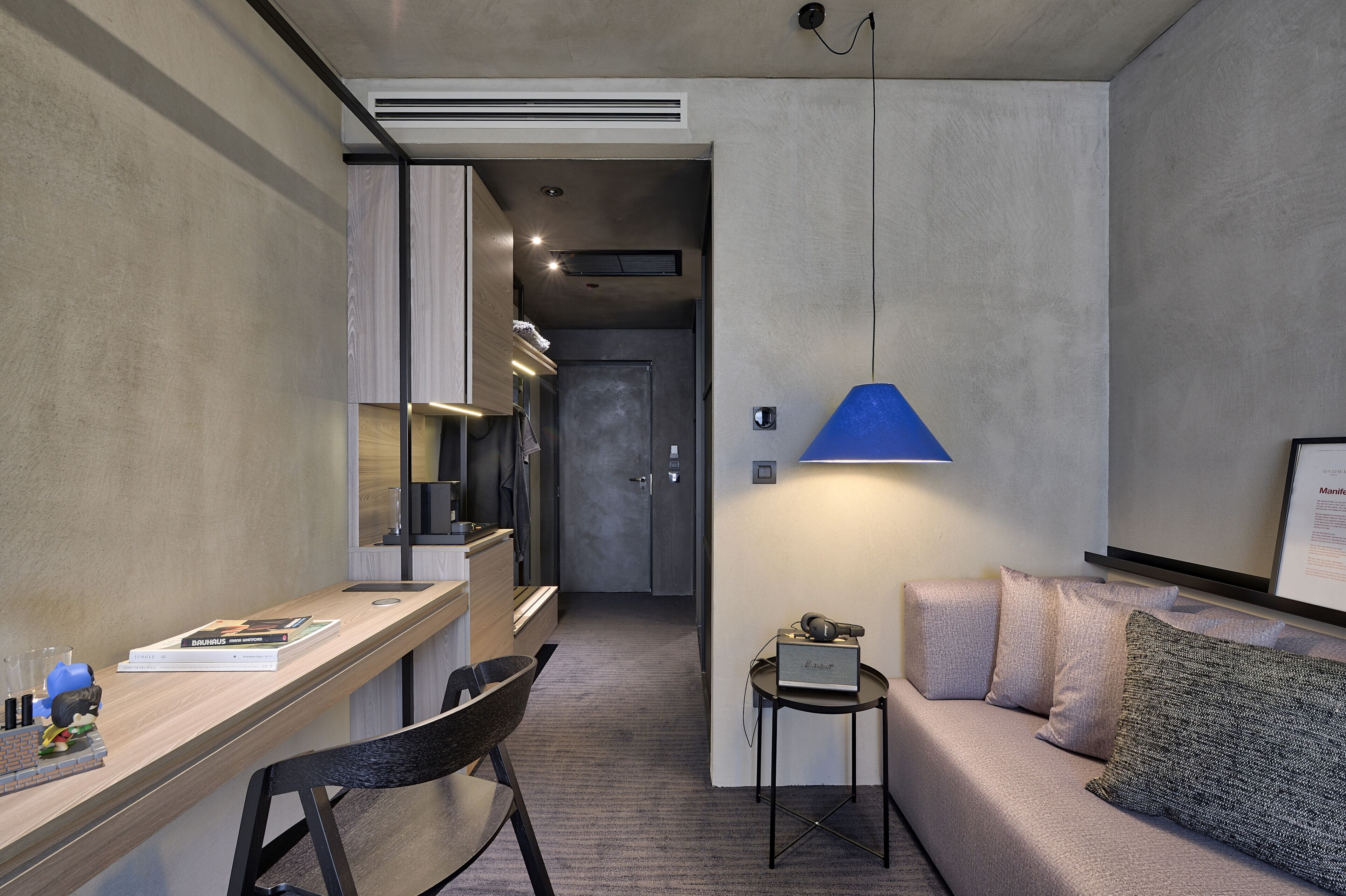 A modern hotel room with a wooden desk, a small table with a lamp, a sofa, and a door leading to a hallway.