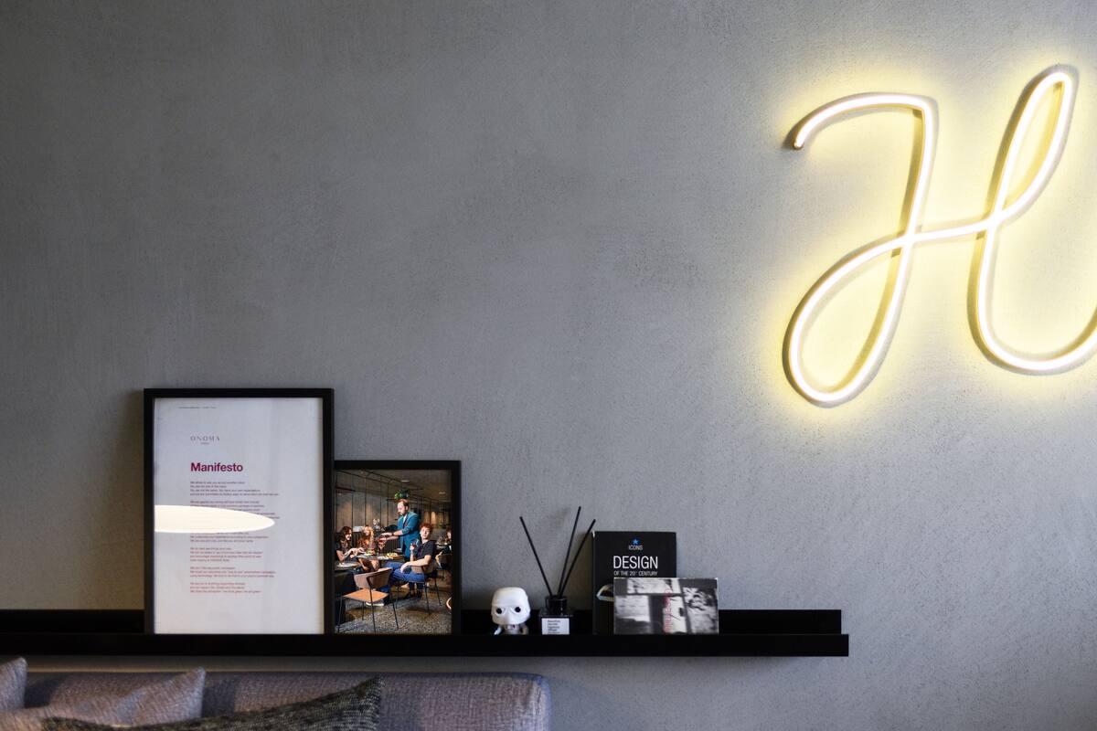 A wall with a neon 'Hi' sign, a framed picture of a restaurant scene, and a shelf with a skull, incense sticks, and a book titled 'Design of the 20th Century'.