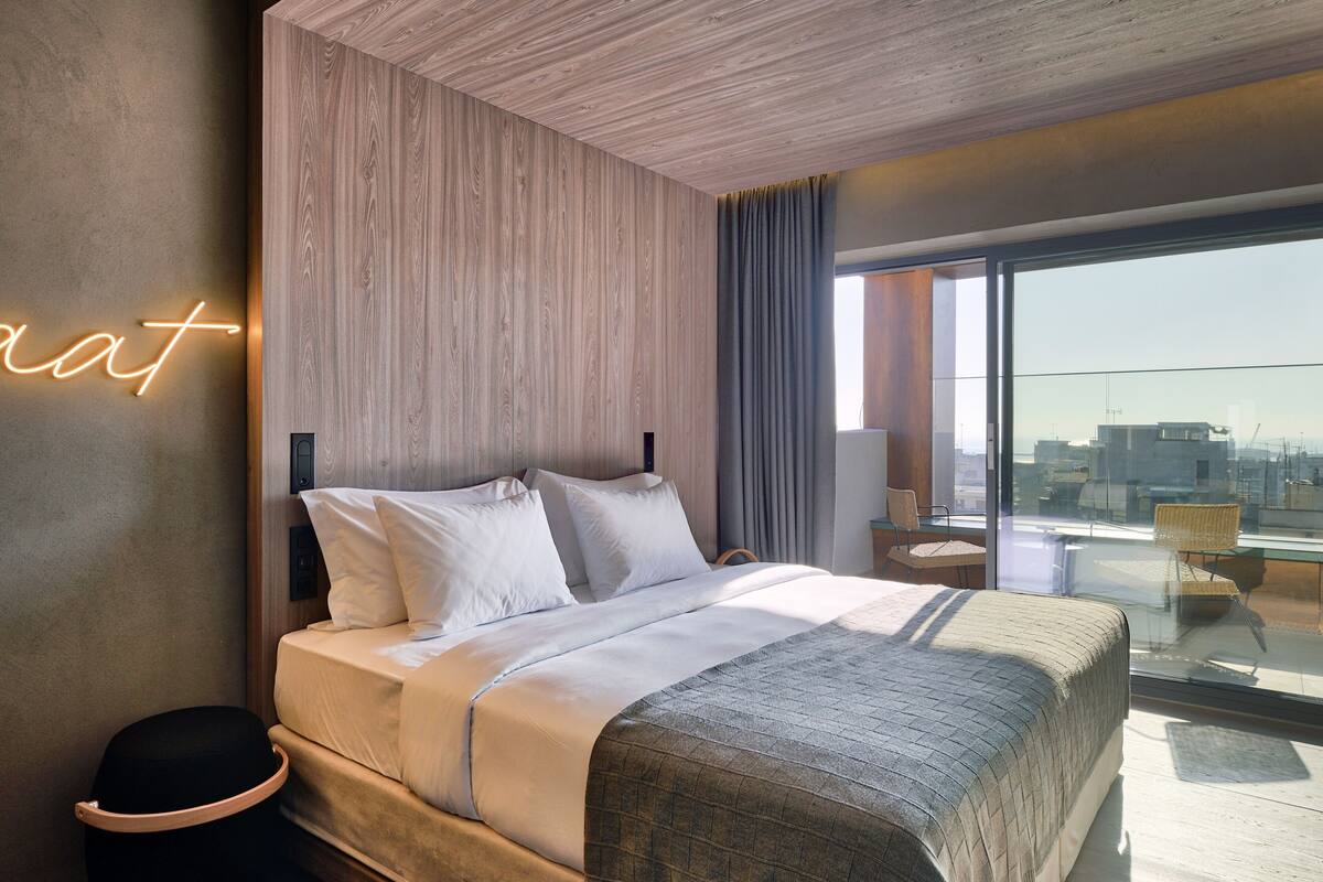 A modern hotel room with a large bed, a wooden accent wall, a neon sign, and a balcony with a view.
