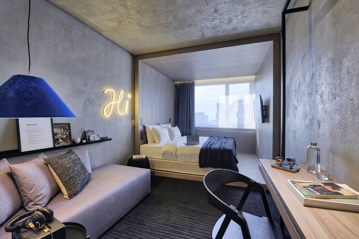 A modern hotel room with a bed, a desk, a chair, a lamp, a neon sign, and a city view.