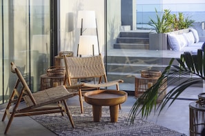 A modern lounge area with wicker furniture, a round wooden table, and a view of the ocean through large glass windows.