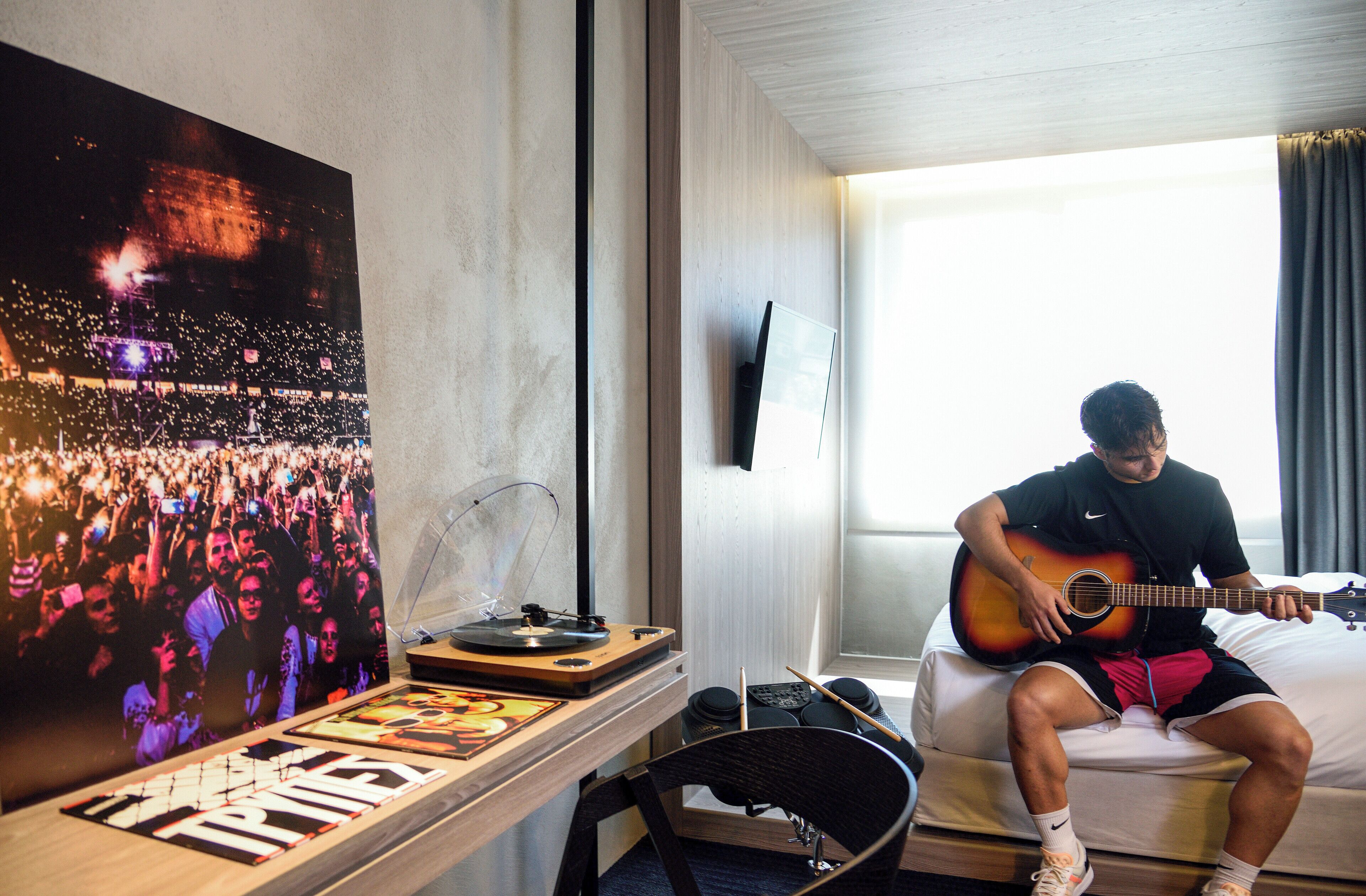 Una habitación con una cama, una persona tocando la guitarra, un cartel y un escritorio con un tocadiscos.