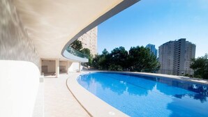 A swimming pool with a clear blue water surface, surrounded by a tiled deck. In the background, there are tall residential buildings and trees. - Apartment Coblanca 16 (Benidorm)