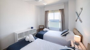 A hotel room with two beds, a ceiling fan, a window with curtains, a small table with a lamp, and a shelf with towels. - Apartment Coblanca 16 (Benidorm)