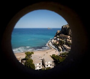 A coastal view through a circular window, featuring a beach, ocean, and buildings on a cliff.