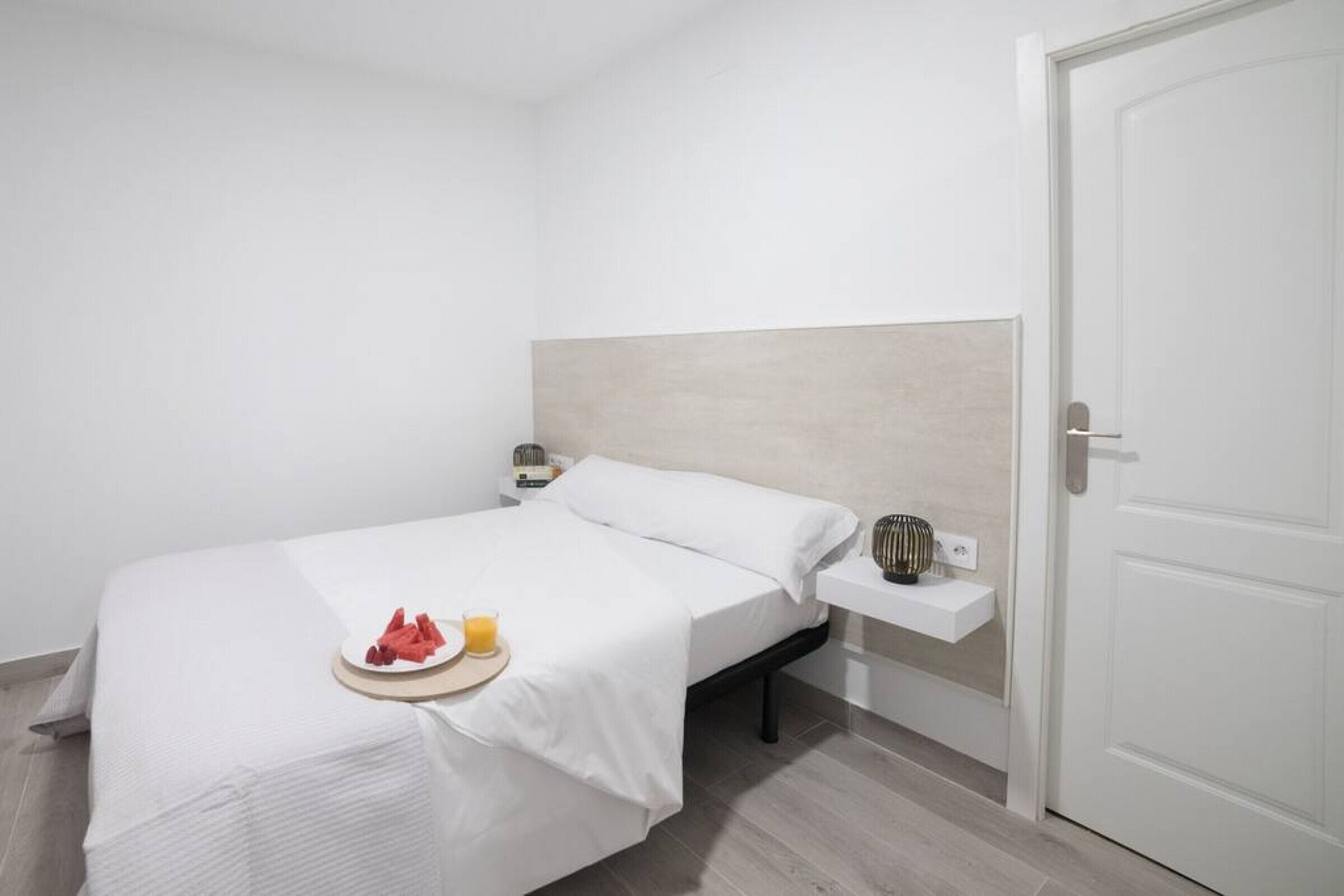 A minimalist hotel room with a bed, a bedside table with a lamp, and a small shelf with a decorative item.