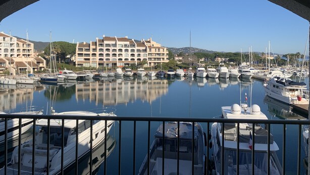 Un port de plaisance où sont amarrés de nombreux bateaux, un grand immeuble doté de balcons et un ciel d’un bleu éclatant.