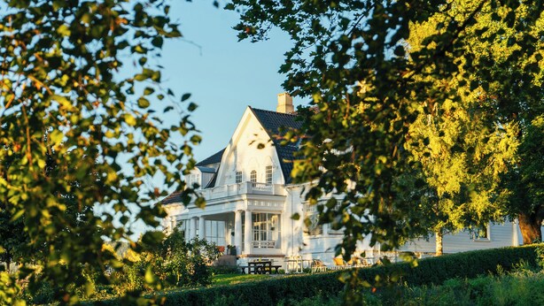 A large, white house with a prominent gable roof, surrounded by lush greenery and trees. - Thorbjørnrud Hotell (Jevnaker)