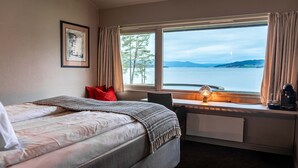A hotel room with a bed, a desk, a window with a view of a lake, and a framed picture on the wall. - Thorbjørnrud Hotell (Jevnaker)
