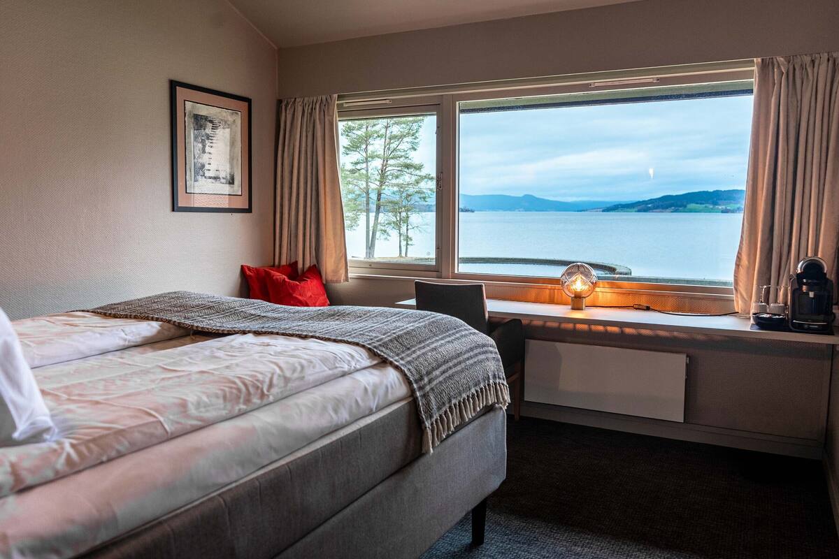 A hotel room with a bed, a desk, a window with a view of a lake, and a framed picture on the wall.