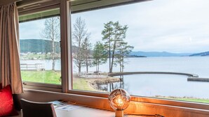 A room with a large window overlooking a lake, a desk with a lamp, and a chair. - Thorbjørnrud Hotell (Jevnaker)