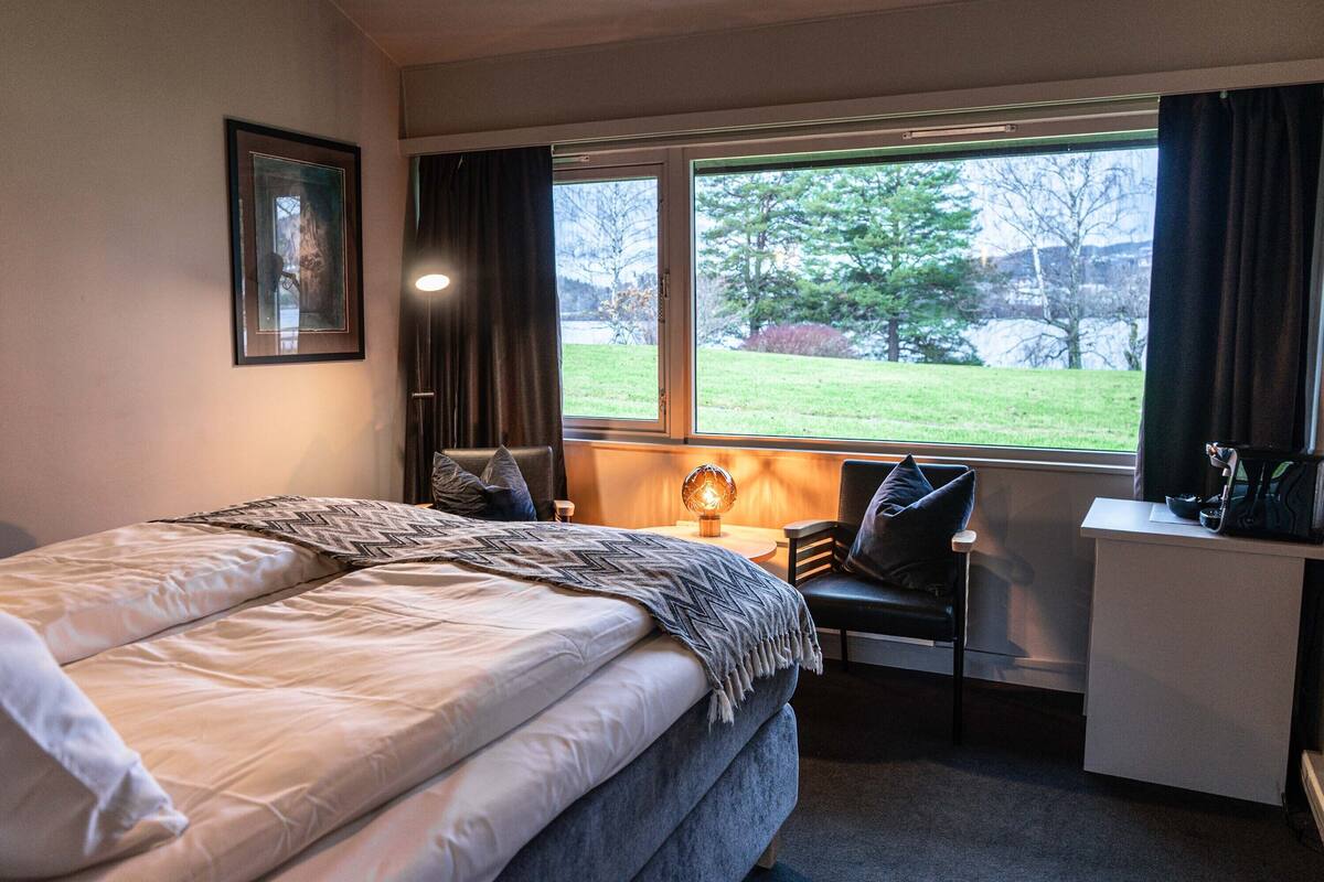 A hotel room with a bed, desk, chair, and a window with a view of a grassy area.