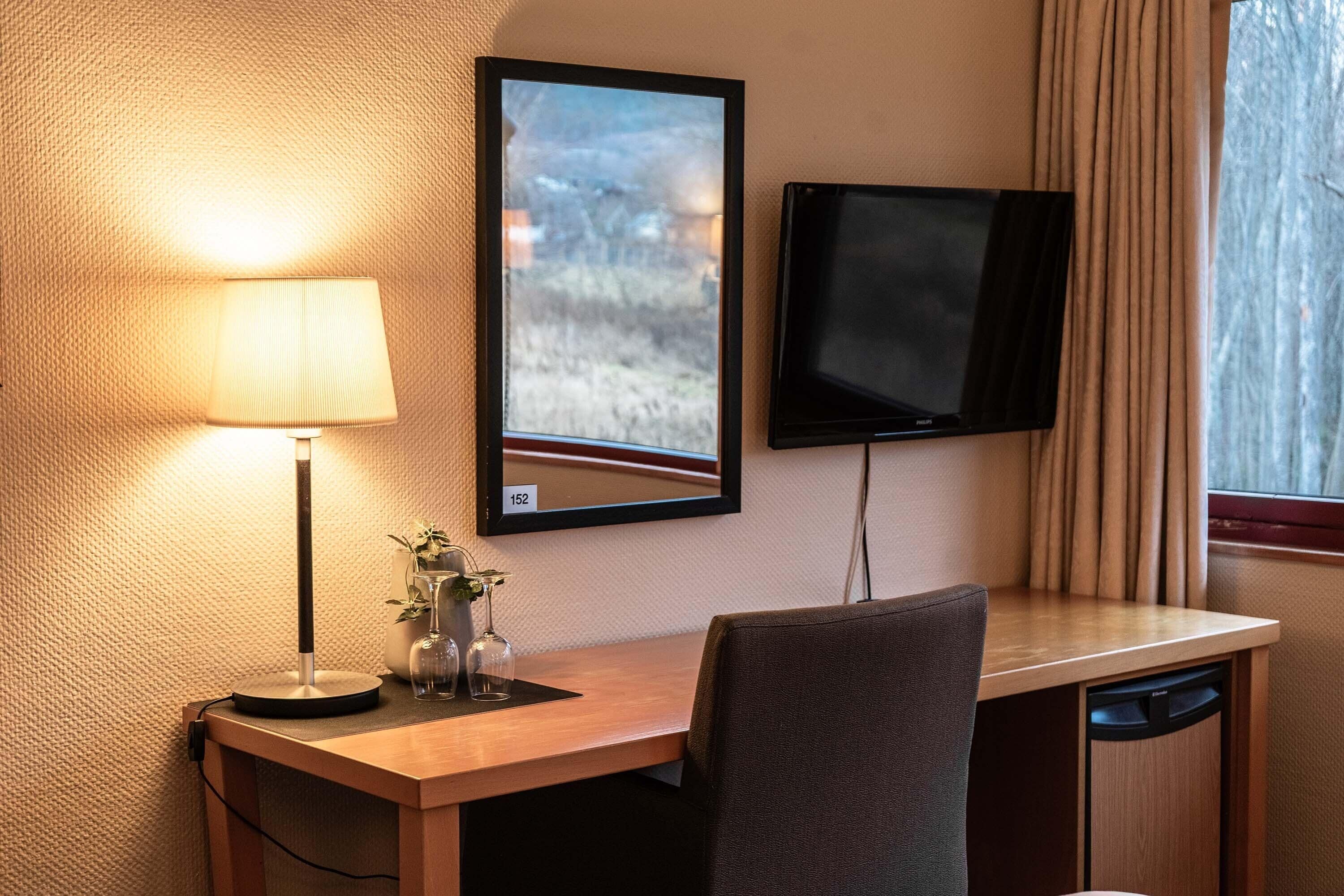 A hotel room with a desk, a lamp, a television, and a window with curtains.