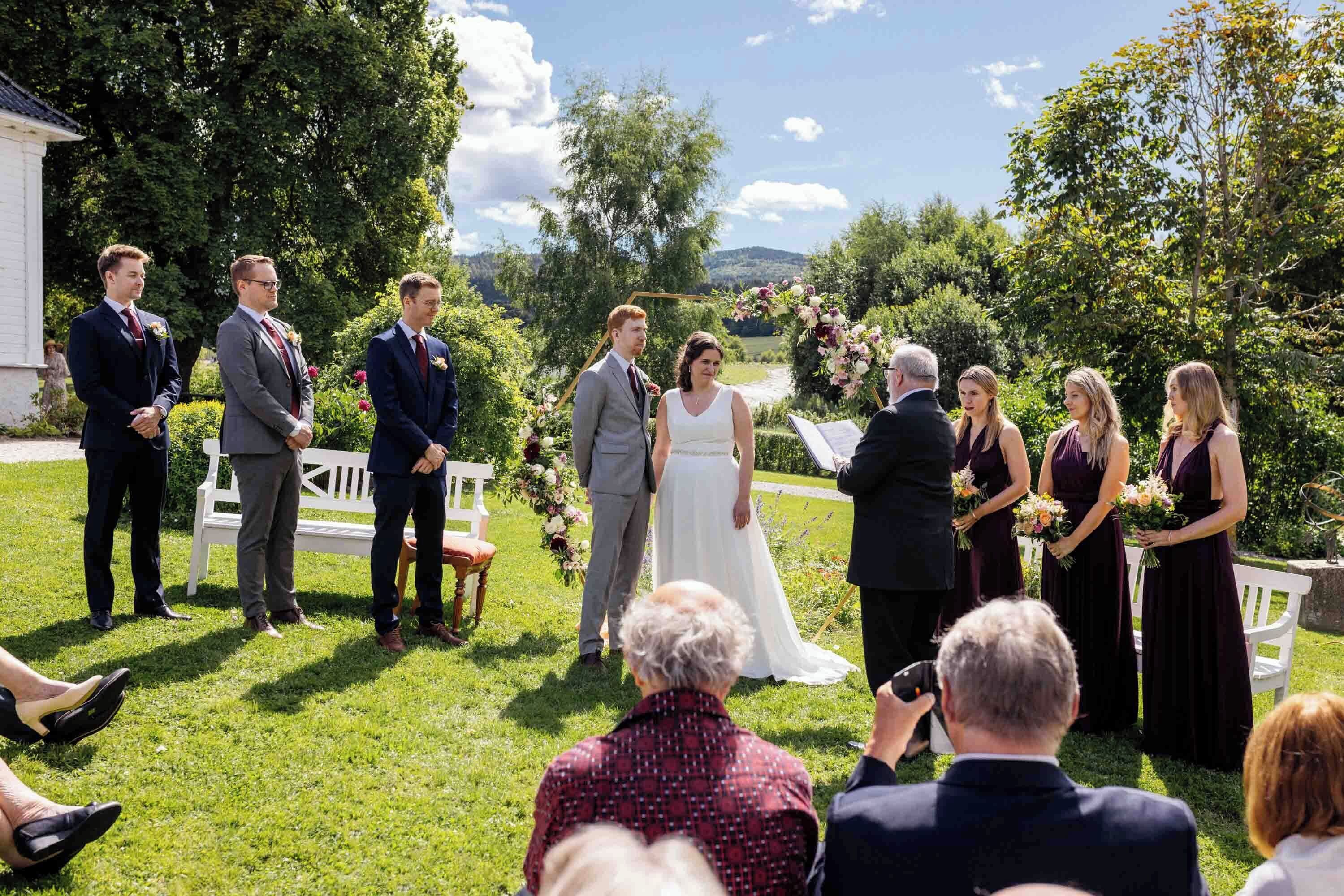 A wedding ceremony outdoors with a grassy area, a white bench, and a backdrop of trees and hills.