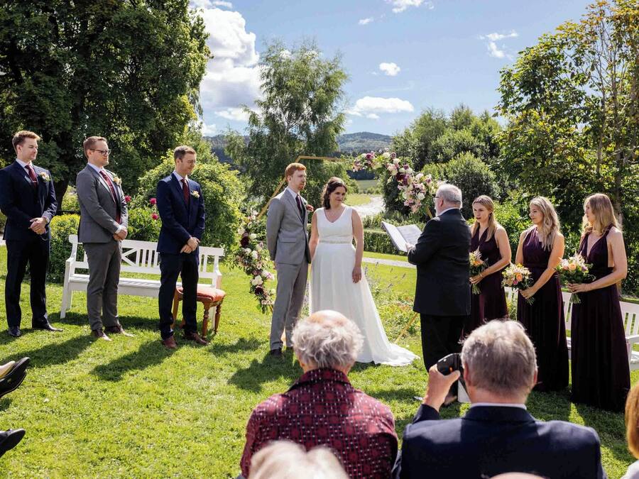 A wedding ceremony outdoors with a grassy area, a white bench, and a backdrop of trees and hills.