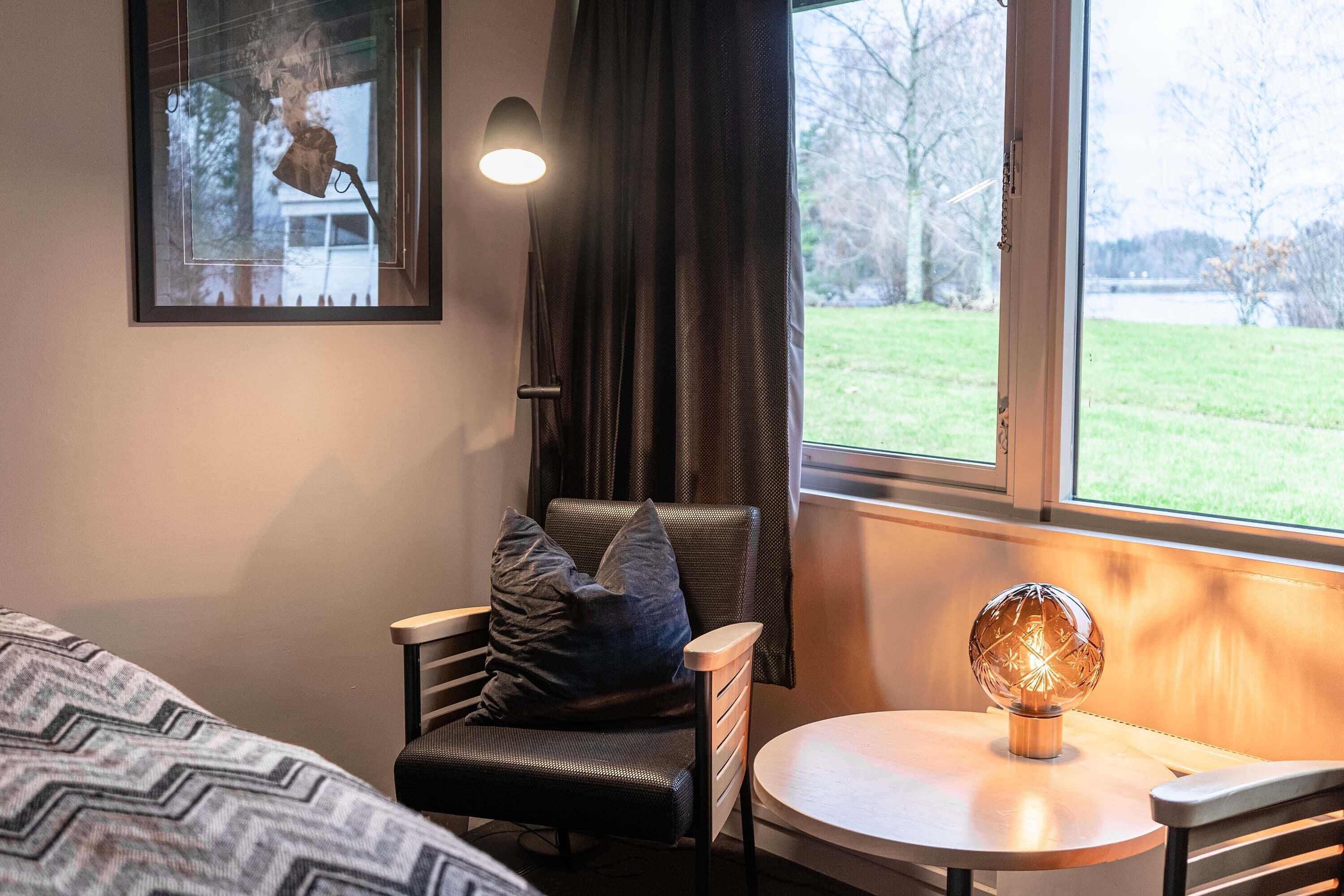 Een hotelkamer met een bed, een stoel, een klein tafeltje, een lamp en een raam met uitzicht op bomen en een gebouw.