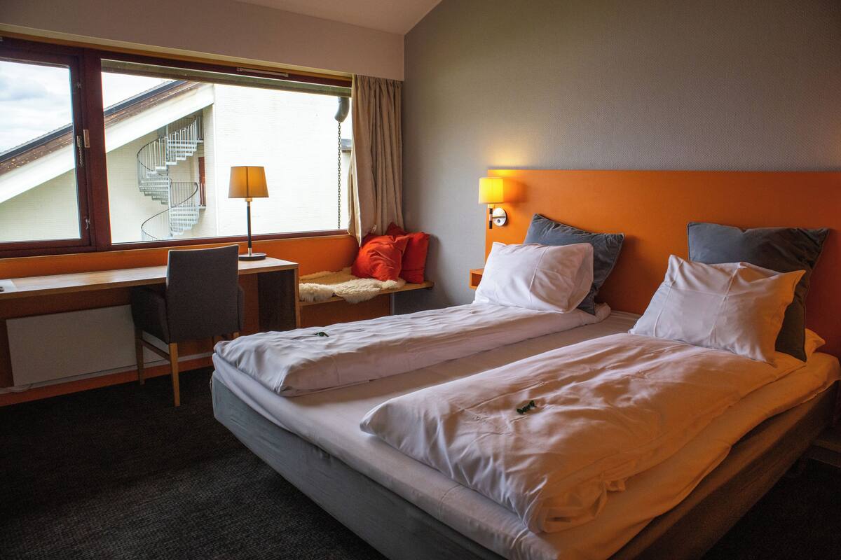 A hotel room with a bed, a desk, a chair, a lamp, and a view of a staircase.