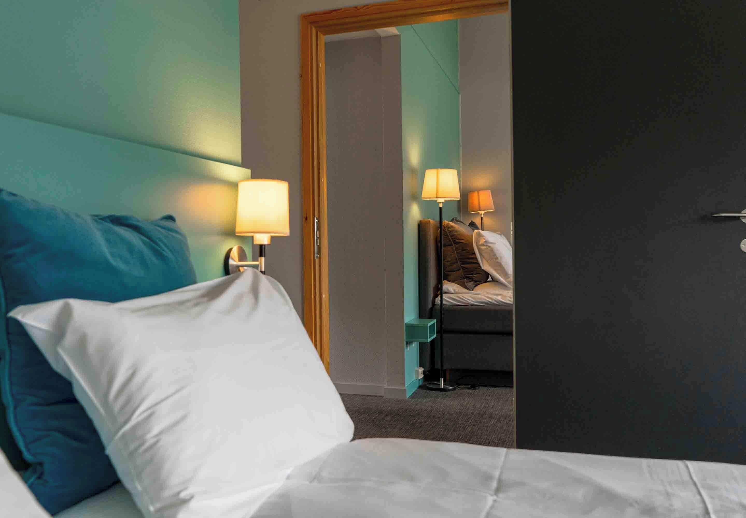 A hotel room with a bed, two bedside lamps, a nightstand, and a door leading to another room.