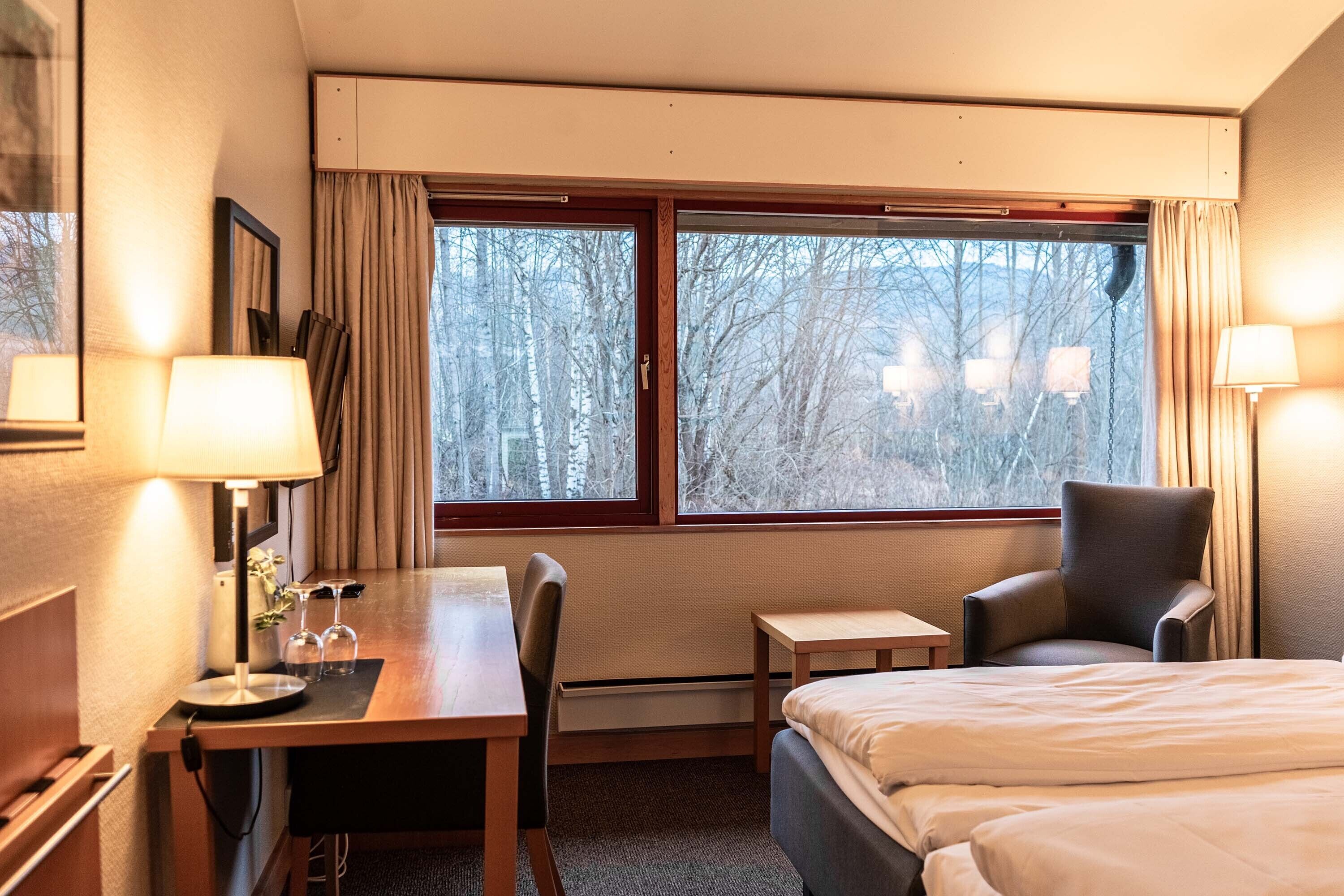 A hotel room with a bed, a desk, a chair, and a window with a view of trees.