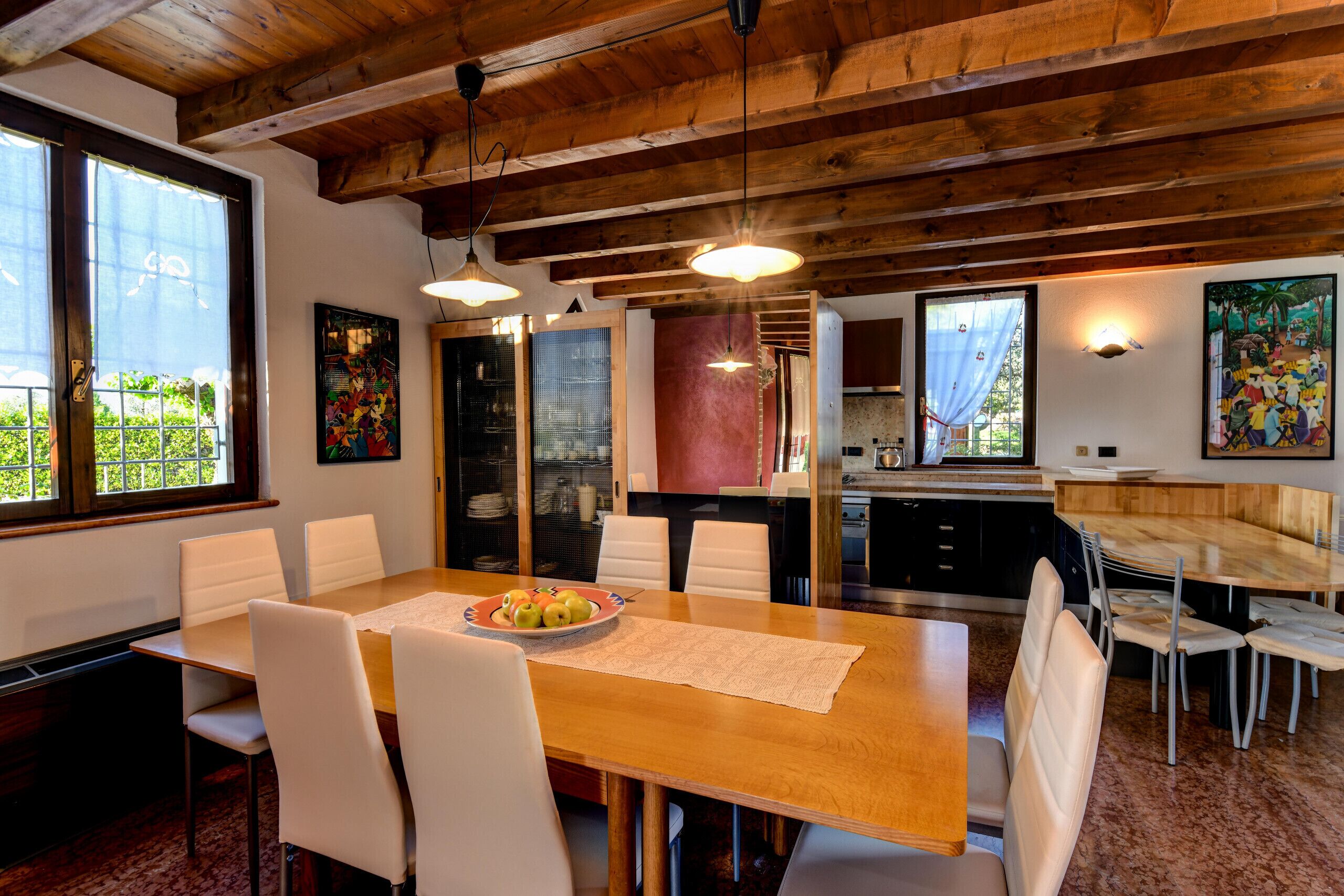 A dining area with a wooden table, white chairs, and a fruit bowl. There are pendant lights above the table, and a kitchen area is visible in the background.