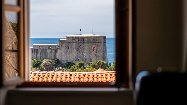 A view of a historic stone building with a flag, through a window with a tiled roof. - Hedera Estate, House Hedera VI (Dubrovnik)