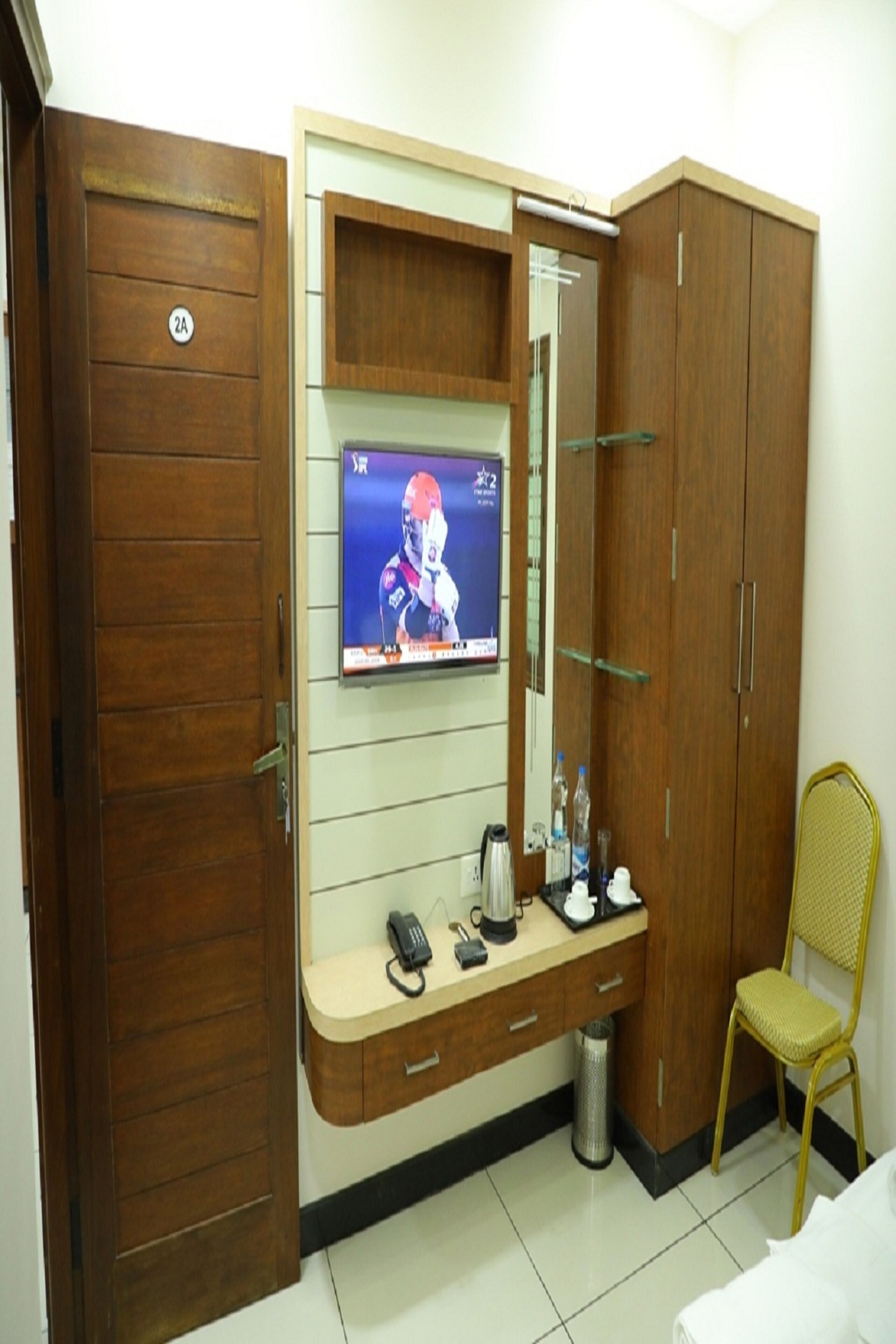 A hotel room with a wooden wardrobe, a television mounted on the wall, a small desk with a telephone, a trash bin, and a yellow chair.