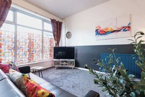 A living room with a sofa, a TV, a plant, and a window with curtains. - Hull Largest 6 Bedrooms House - Sleeps 13 (Hull)