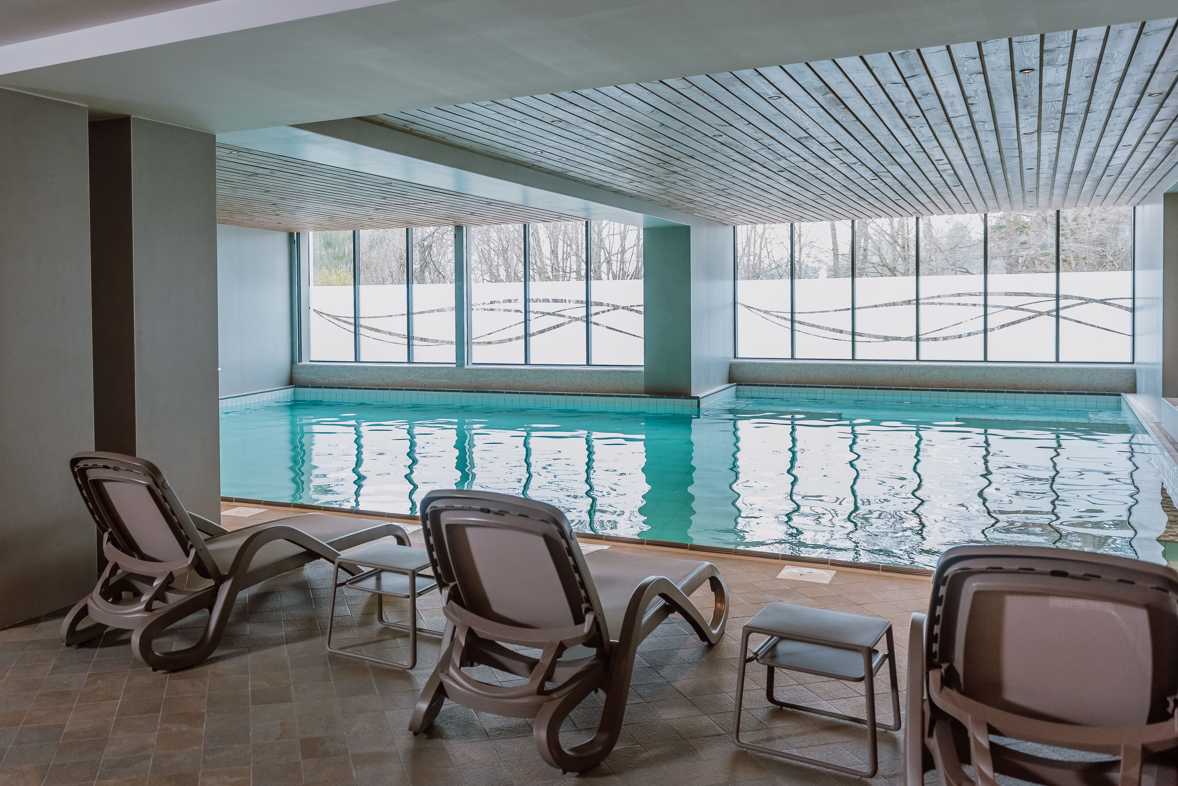 An indoor swimming pool with clear glass walls, several lounge chairs, and a tiled floor.