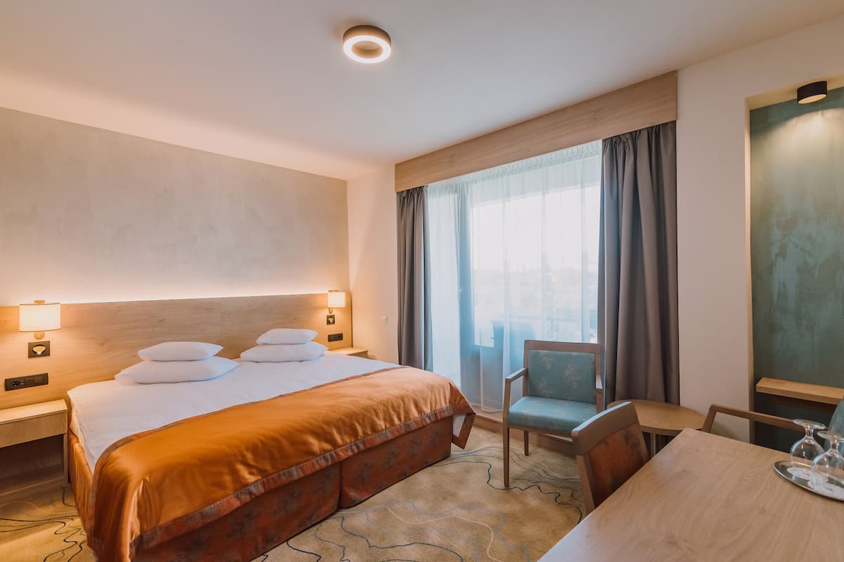 Habitación de hotel con una cama grande, dos lámparas de noche, cabecera de madera, una ventana con cortinas, una silla y una mesa con bandeja.