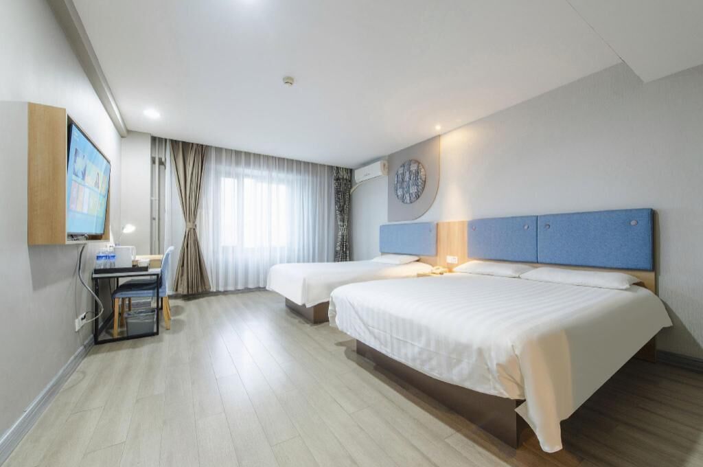 A hotel room with two beds, a desk with a computer, a TV, and a large window with curtains.