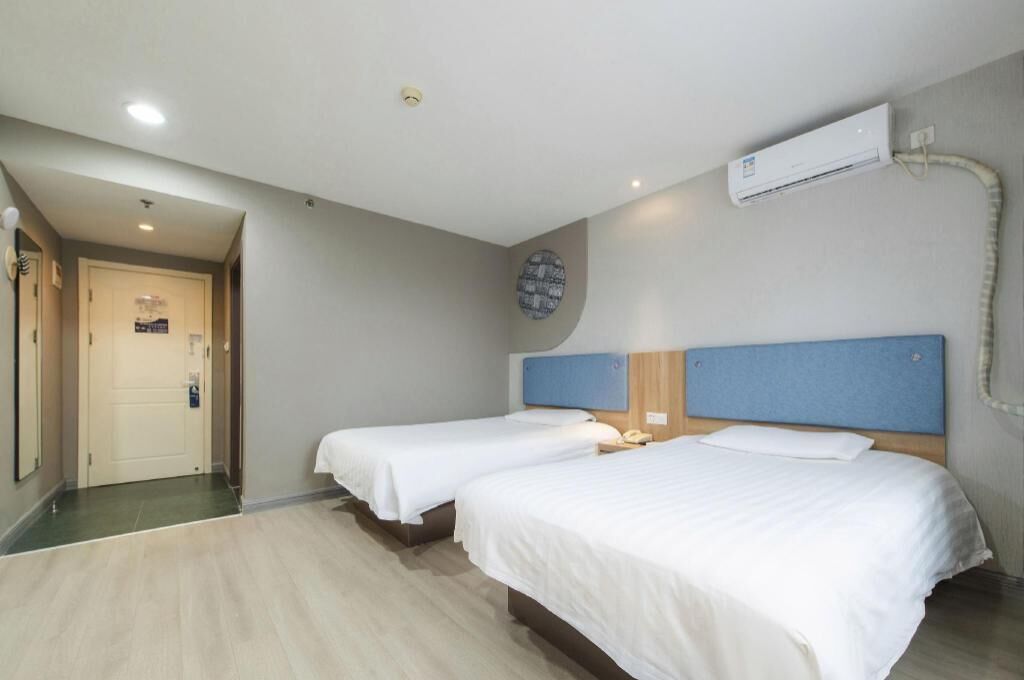 Une chambre d’hôtel avec deux lits simples, un ventilateur de plafond, un appareil de climatisation et une petite horloge ronde fixée au mur.
