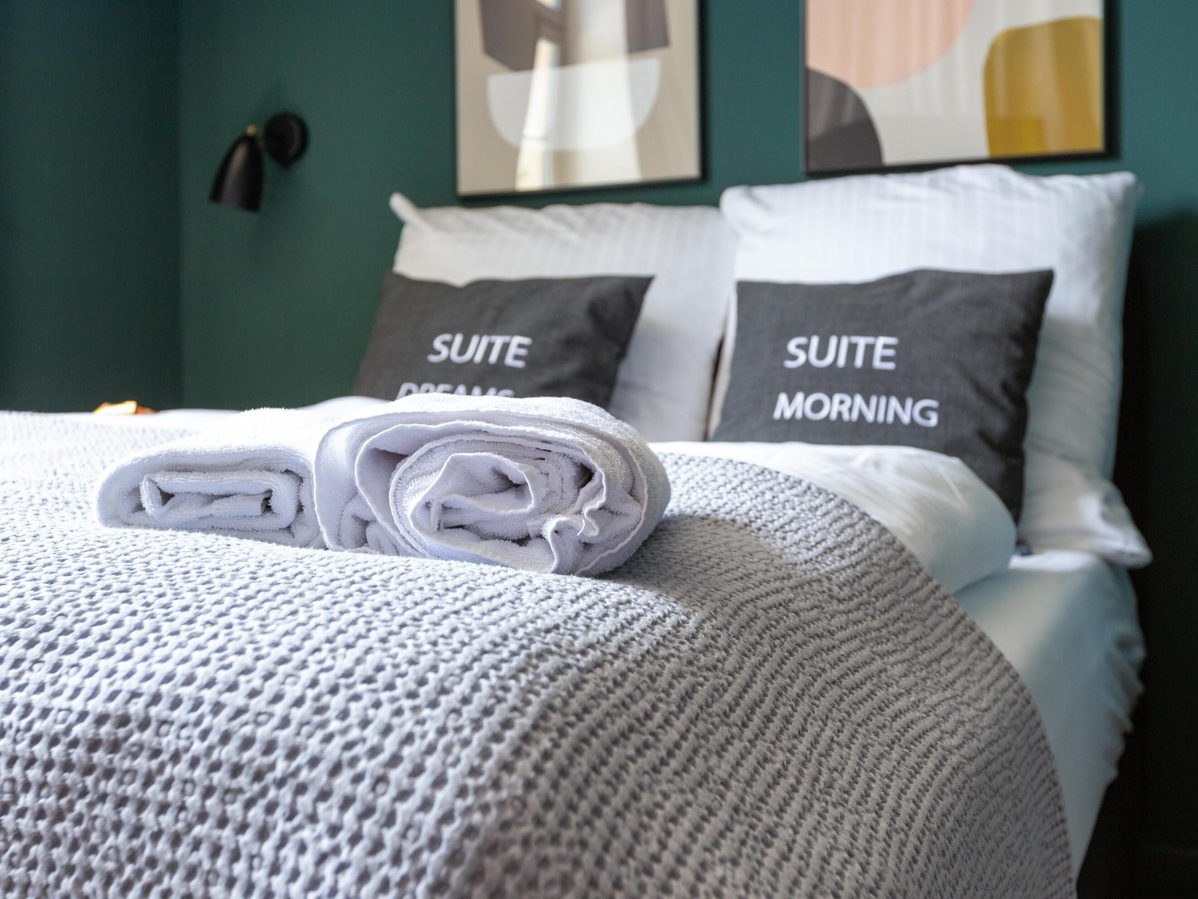 Un lit bien fait, avec une serviette blanche roulée et des oreillers portant les inscriptions « RÊVES DE SUITE » et « MATIN DE SUITE », dans une pièce aux murs de couleur turquoise et décorée d’œuvres d’art abstraites.