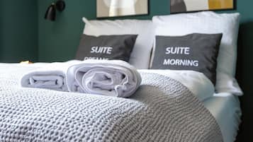 Un lit bien fait, avec une serviette blanche roulée et des oreillers portant les inscriptions « RÊVES DE SUITE » et « MATIN DE SUITE », dans une pièce aux murs de couleur turquoise et décorée d’œuvres d’art abstraites.