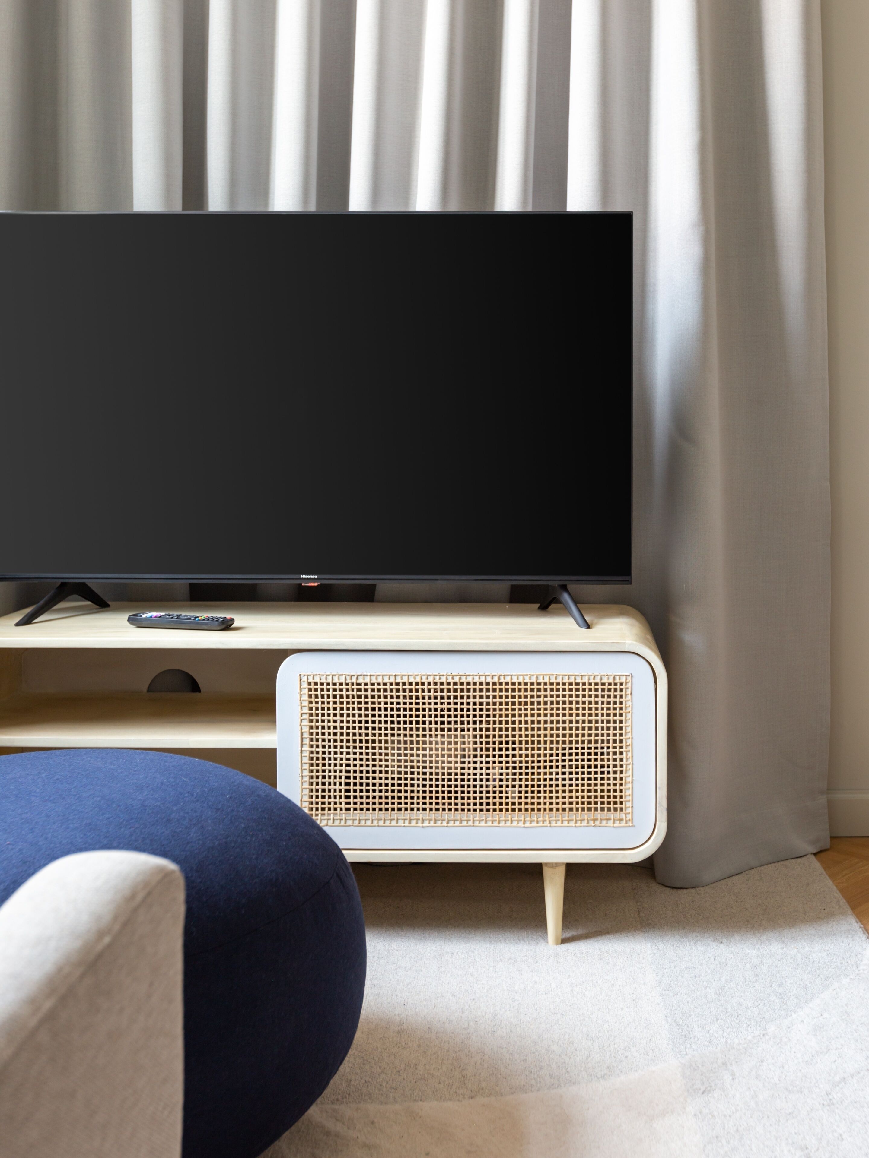 Un televisor de pantalla plana sobre un soporte de madera, con un altavoz tejido debajo, ubicado frente a una cortina.