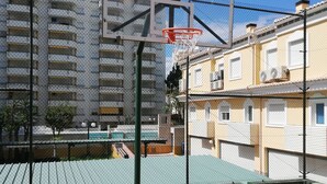 An outdoor basketball court with a net and backboard, surrounded by a chain-link fence, with residential buildings in the background. - CANCUN VII 6º,  Solo Familias! (Playa de Gandía)