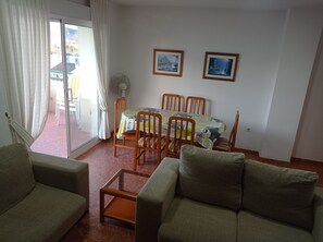 A living room with a sofa, a glass coffee table, a dining table with chairs, a fan, and two framed pictures on the wall.