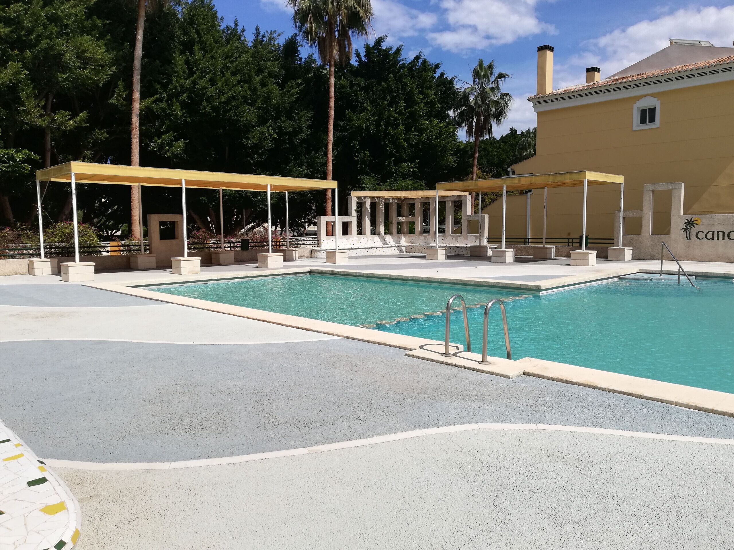 Una piscina con zona techada, palmeras y un edificio con un logotipo.