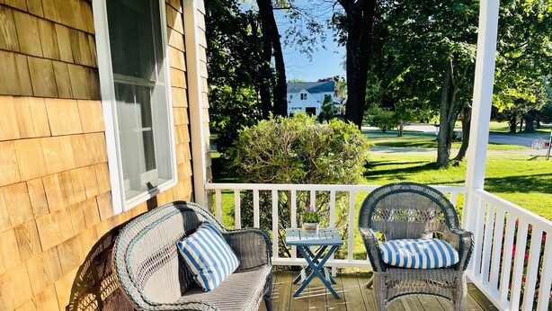 En veranda av trä med möbler av rotting och ett litet bord, med utsikt över en grön gräsmatta och ett hus i bakgrunden.