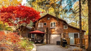 Una cabaña rústica con un garaje, un patio con mobiliario exterior y un árbol de hojas rojas cerca.