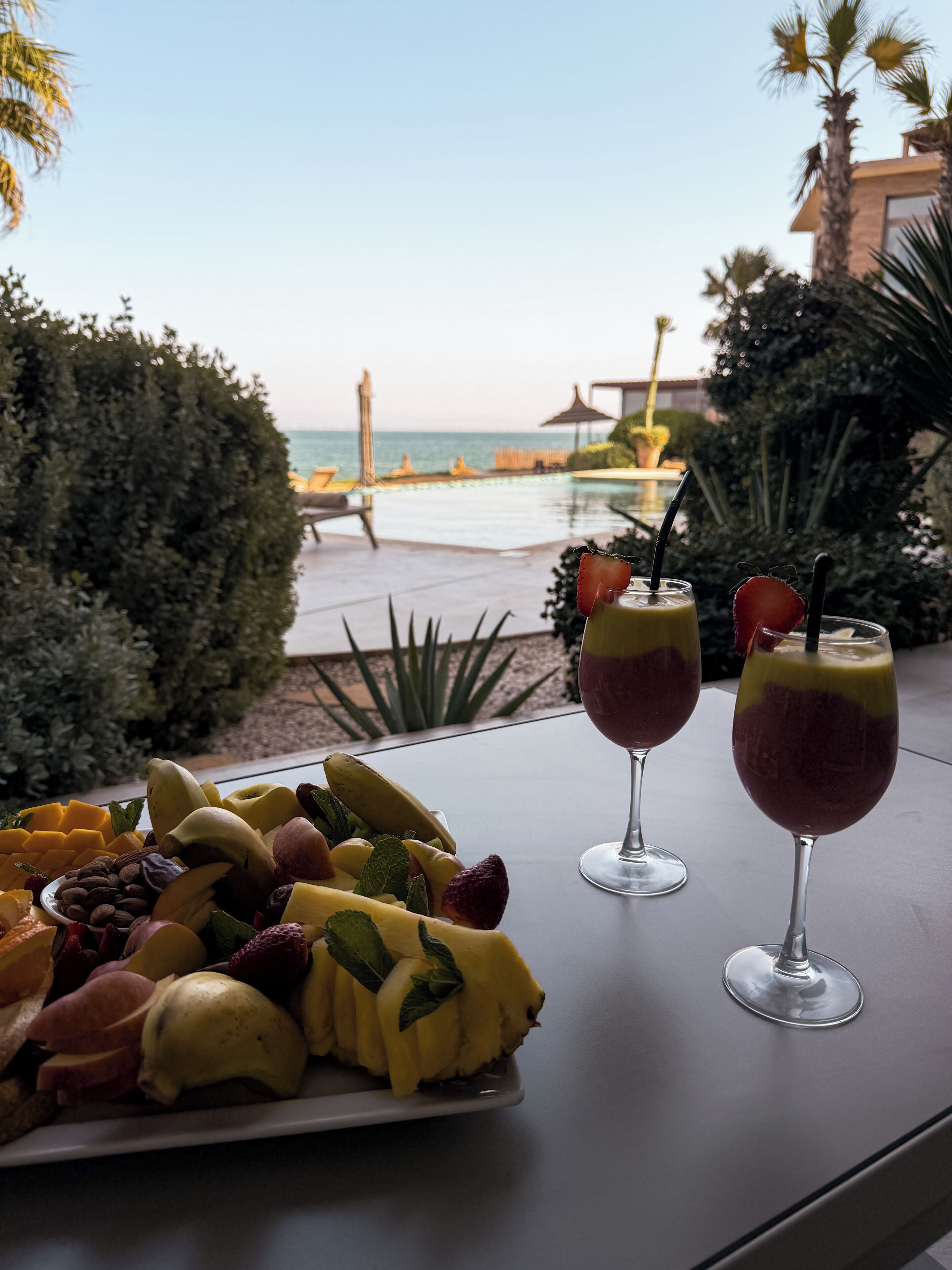 A table with a fruit platter and two drinks, overlooking a pool and ocean view.
