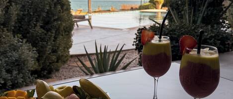 A table with a fruit platter and two drinks, overlooking a pool and ocean view.