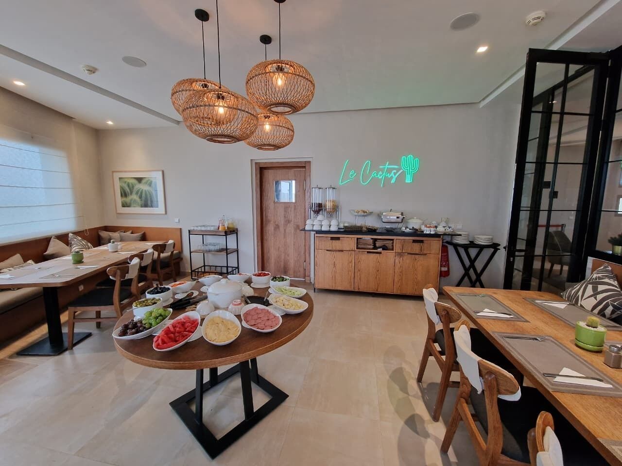 A modern dining area with a round table set for a meal, a kitchen in the background, and a neon sign on the wall.