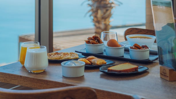 A breakfast table with food and drinks, a window with a view of palm trees and the sea.