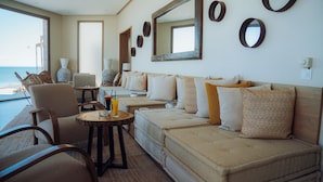 A room with a sofa, chairs, a table with drinks, and a large window overlooking the sea.